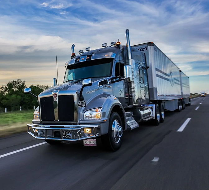 Camión Kenworth plateado transportando un remolque blanco en carretera bajo cielo azul con nubes.