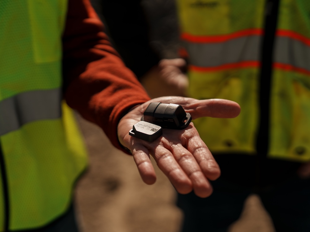 Trabajador con chaleco reflectante sosteniendo pequeños dispositivos electrónicos en su mano, posiblemente sensores o rastreadores.