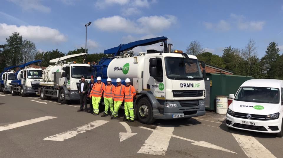 Five Drainline workers in orange vests and hard hats stand beside a fleet of white Drainline service trucks in a parking lot.