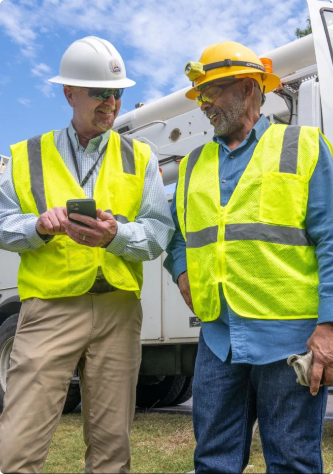 Two construction workers in safety vests and hard hats looking at a smartphone next to a utility truck.