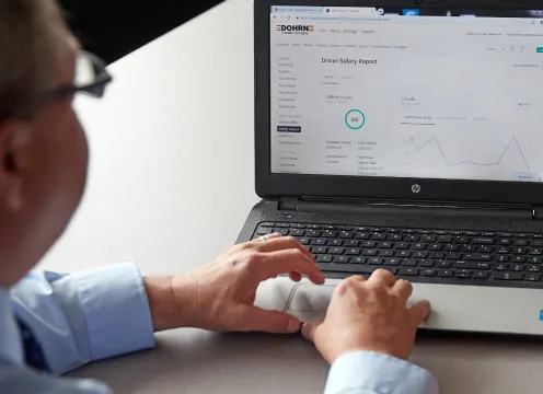 A seated person looking at a laptop on a desk with their hands on the keyboard with another person looking on