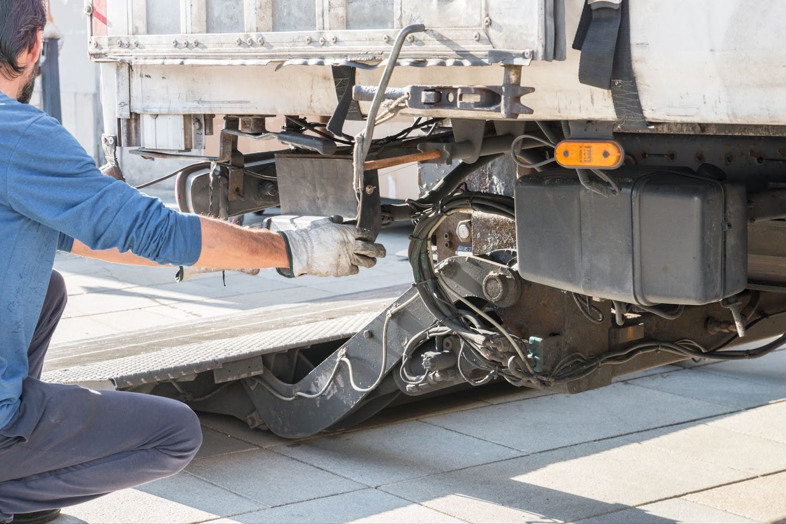 Mechanic in blue shirt and work gloves repairing the undercarriage of a truck or trailer with exposed mechanical components.