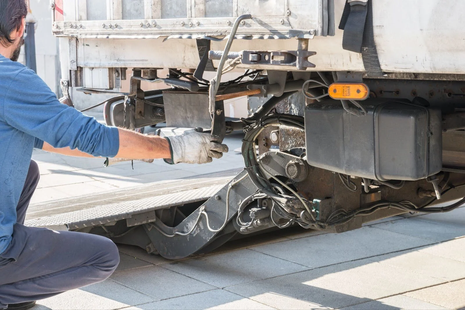 Mechanic in blue shirt and work gloves repairing the undercarriage of a truck or trailer with exposed mechanical components.