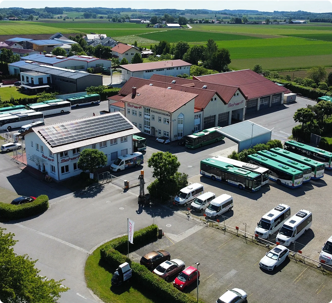 Aerial view of a bus depot with red-roofed buildings, parked buses, and surrounding rural farmland with green fields.