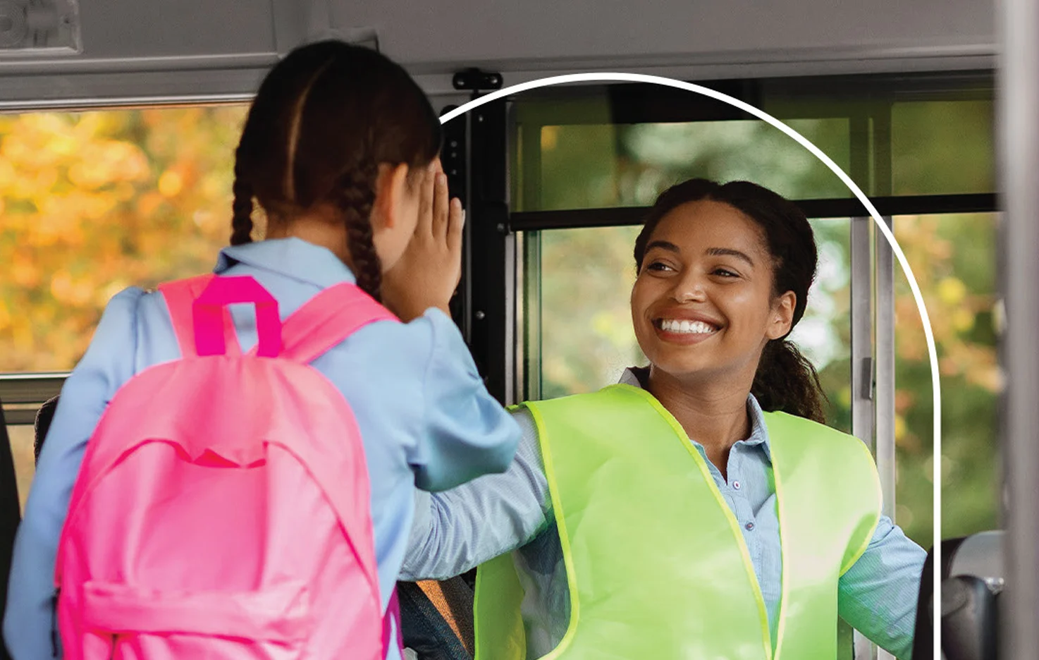 A woman in a safety vest smiles at a child with a pink backpack on a school bus.