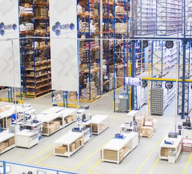 Large warehouse interior with blue storage racks filled with boxes and workstations with wooden crates on the floor.