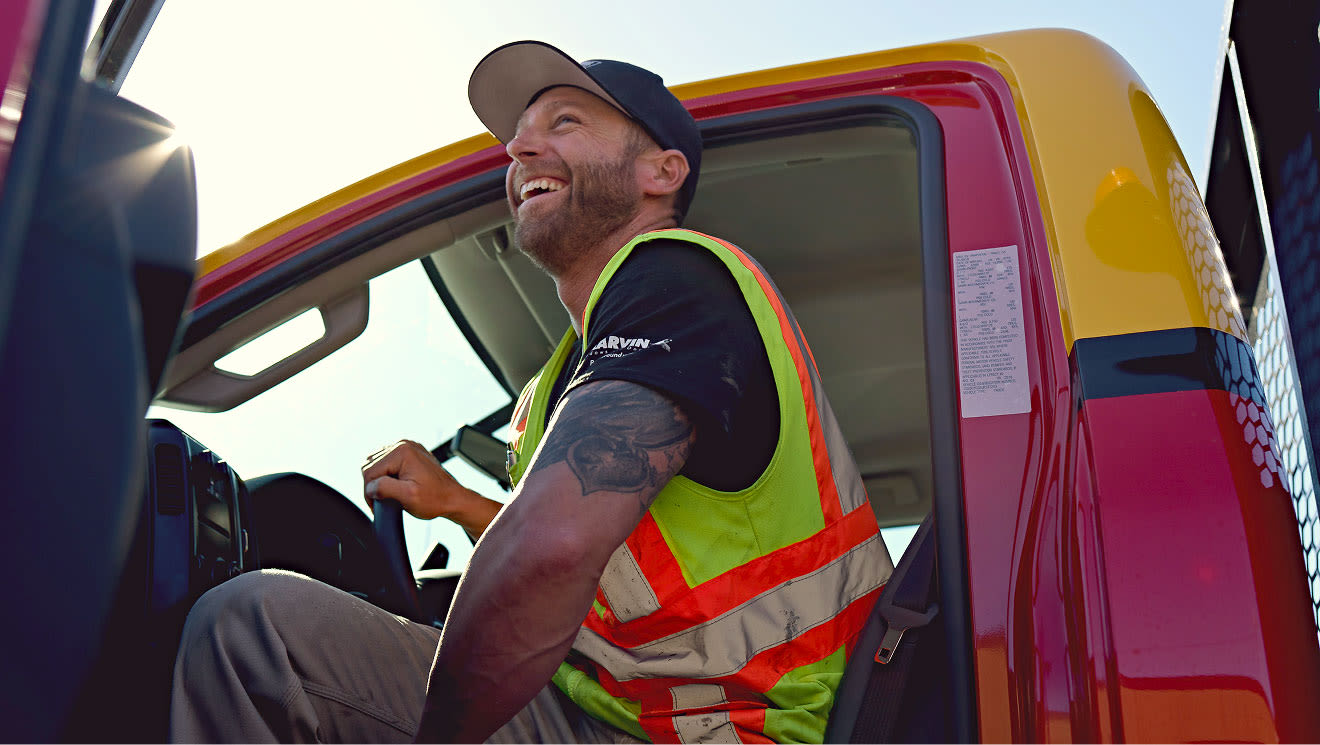 A smiling man in a yellow safety vest and cap sits in a red and yellow truck cab, laughing outdoors.