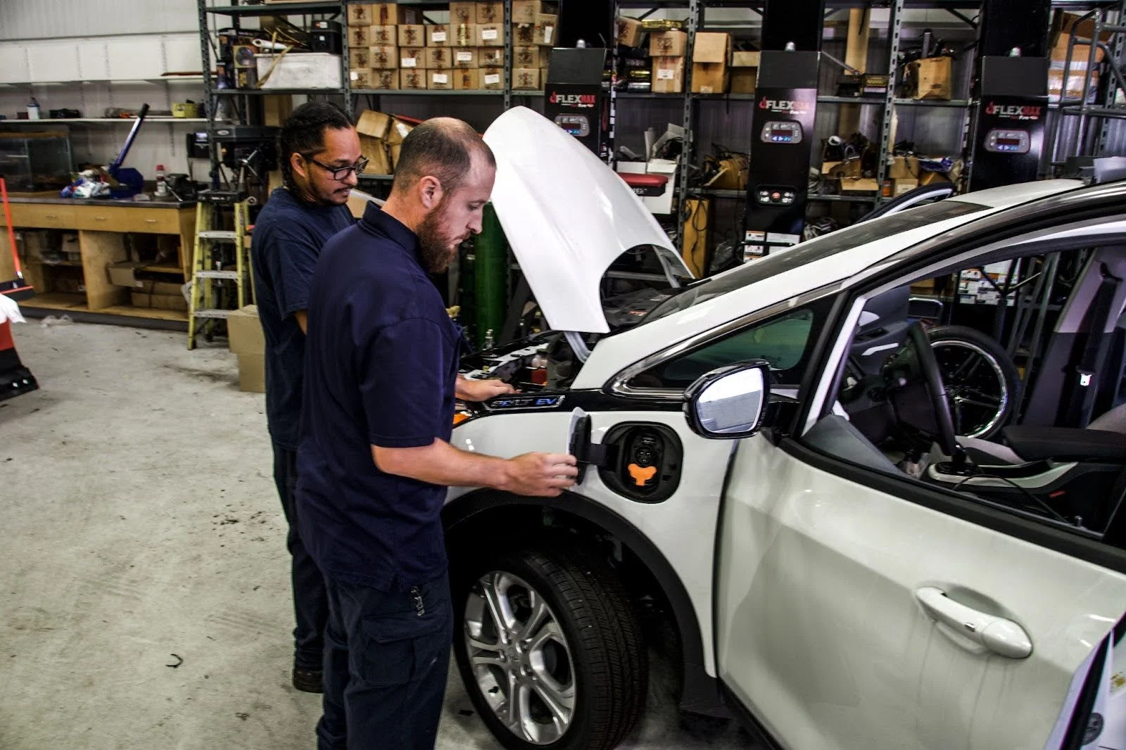 Two mechanics working on a white electric vehicle in an auto repair shop with storage shelves in the background.