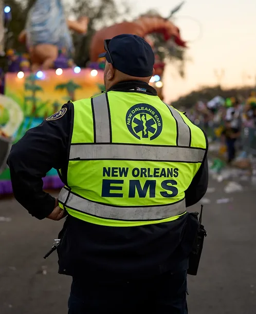 New Orleans EMS worker in bright yellow safety vest watching over a parade with colorful floats in background.