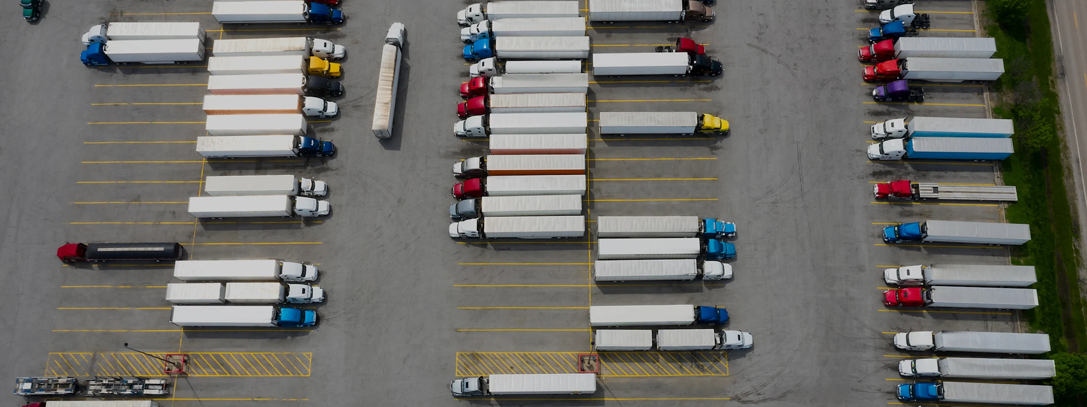 Aerial view of a truck parking lot with numerous semi-trucks with white trailers parked in organized rows with yellow lines.