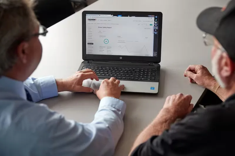 Two men reviewing a Driver Safety Report on an HP laptop, viewed from above at a table.