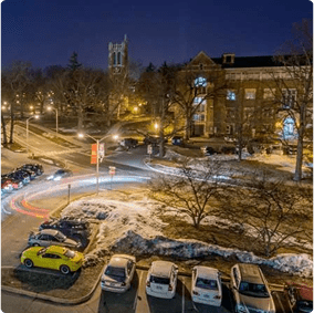 Nighttime campus view with illuminated buildings, clock tower, parked cars, and snow-covered grounds.