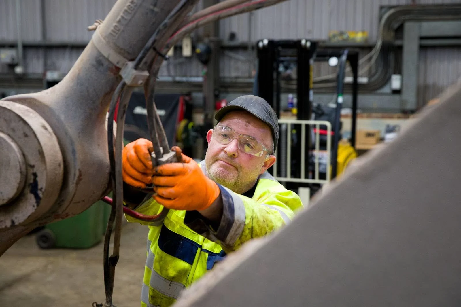 Industrial worker in safety vest and orange gloves repairing machinery in a factory workshop.