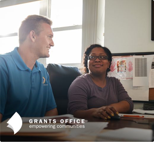 Two people in conversation at a desk in the Grants Office with "empowering communities" tagline below.