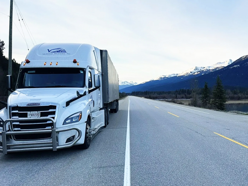 White semi truck driving on a highway with snow-capped mountains in the background under clear blue sky.