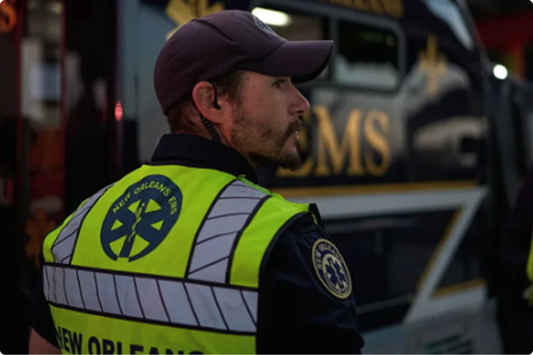 EMS worker in neon yellow safety vest and purple cap standing beside an ambulance in New Orleans at night.