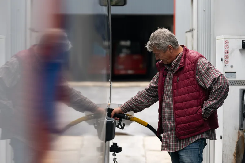 Un homme âgé en gilet rouge ravitaille un véhicule avec un pistolet à carburant jaune dans une station-service.