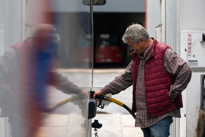 Un homme âgé en gilet rouge ravitaille un véhicule avec un pistolet à carburant jaune dans une station-service.
