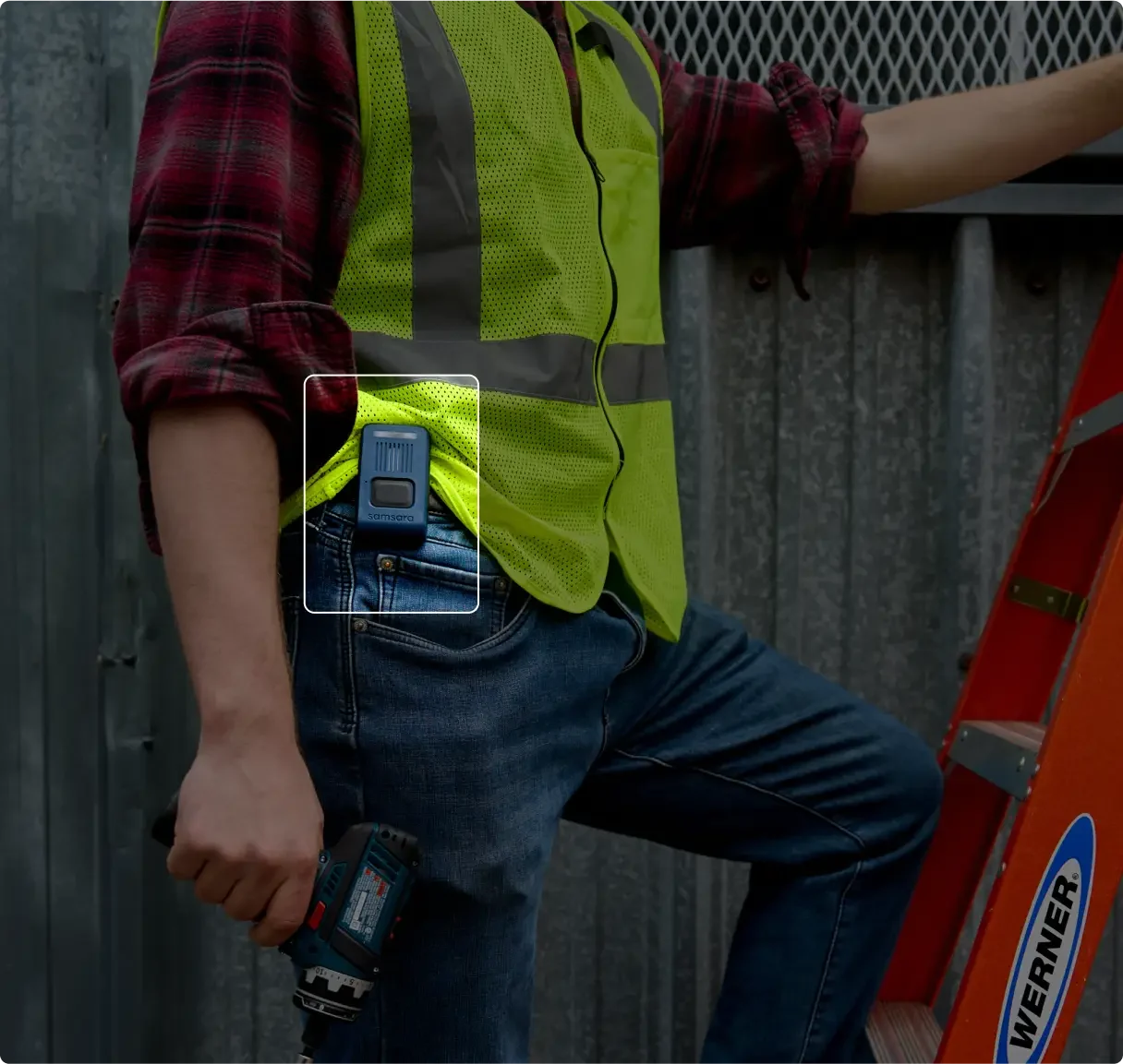 Construction worker in hi-vis vest and plaid shirt holding drill, with Samsara wearable clipped to jeans pocket.