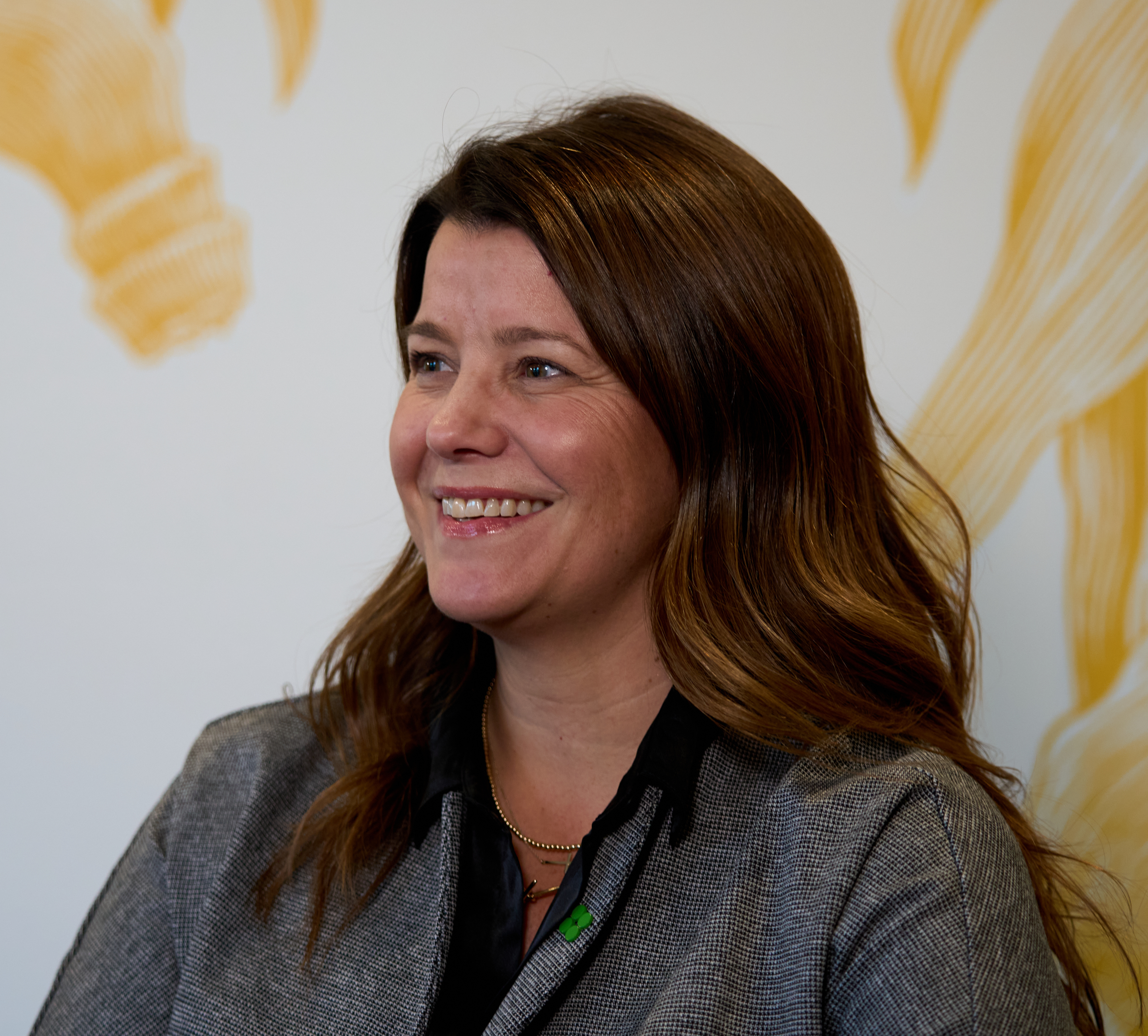 Woman with brown hair smiling, wearing gray blazer and black top against white background with gold accents.