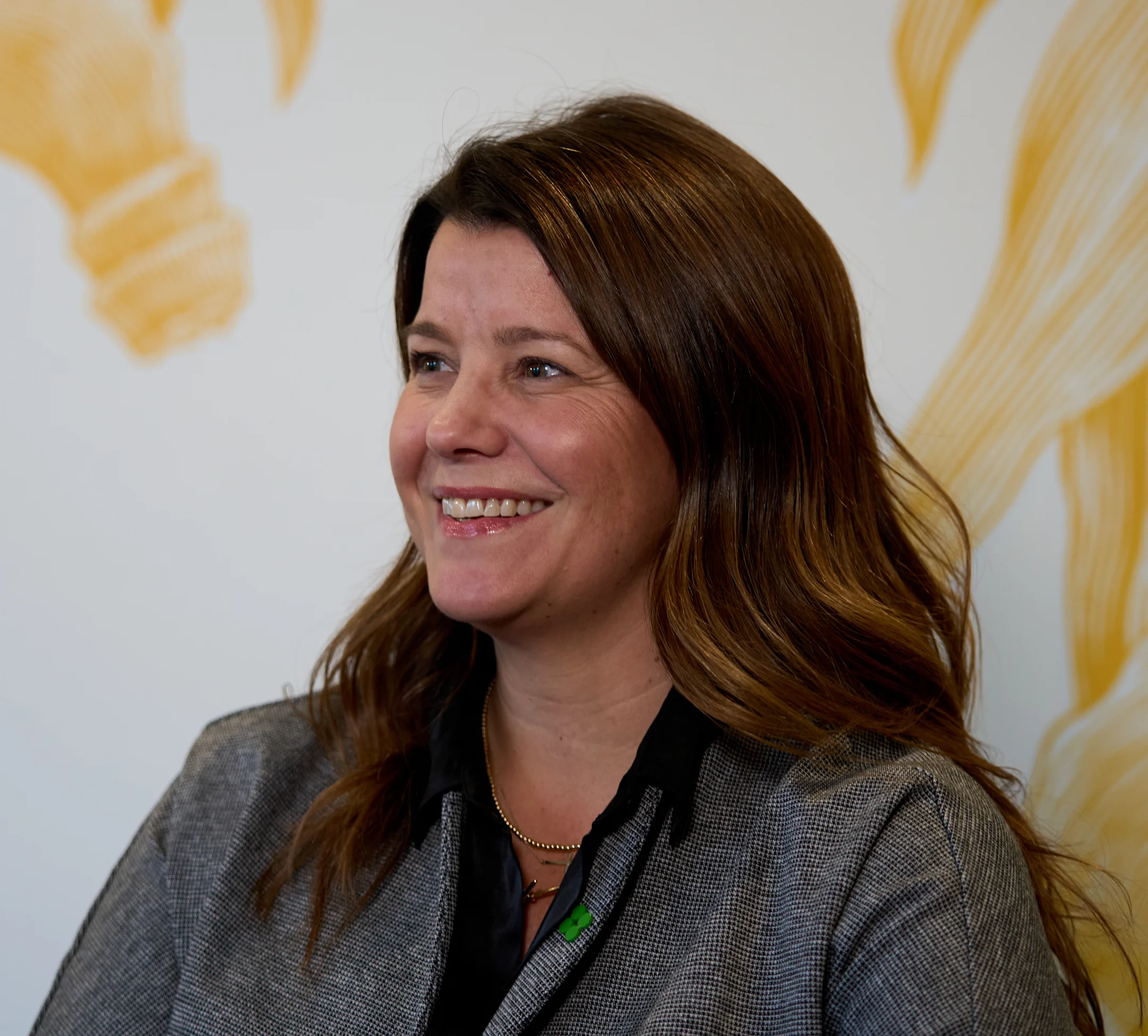 Woman with brown hair smiling, wearing gray blazer and black top against white background with gold accents.