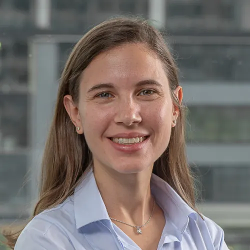 Mujer sonriente con cabello castaño y camisa azul clara en un entorno de oficina con fondo de edificio moderno.