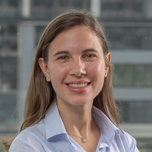 A smiling person with long brown hair wearing a light blue button-up shirt against a blurred office building background.