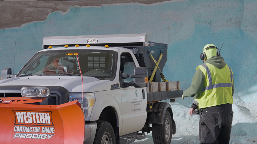 Worker in yellow vest guides a white truck with a Western Prodigy snow plow at a salt storage facility.