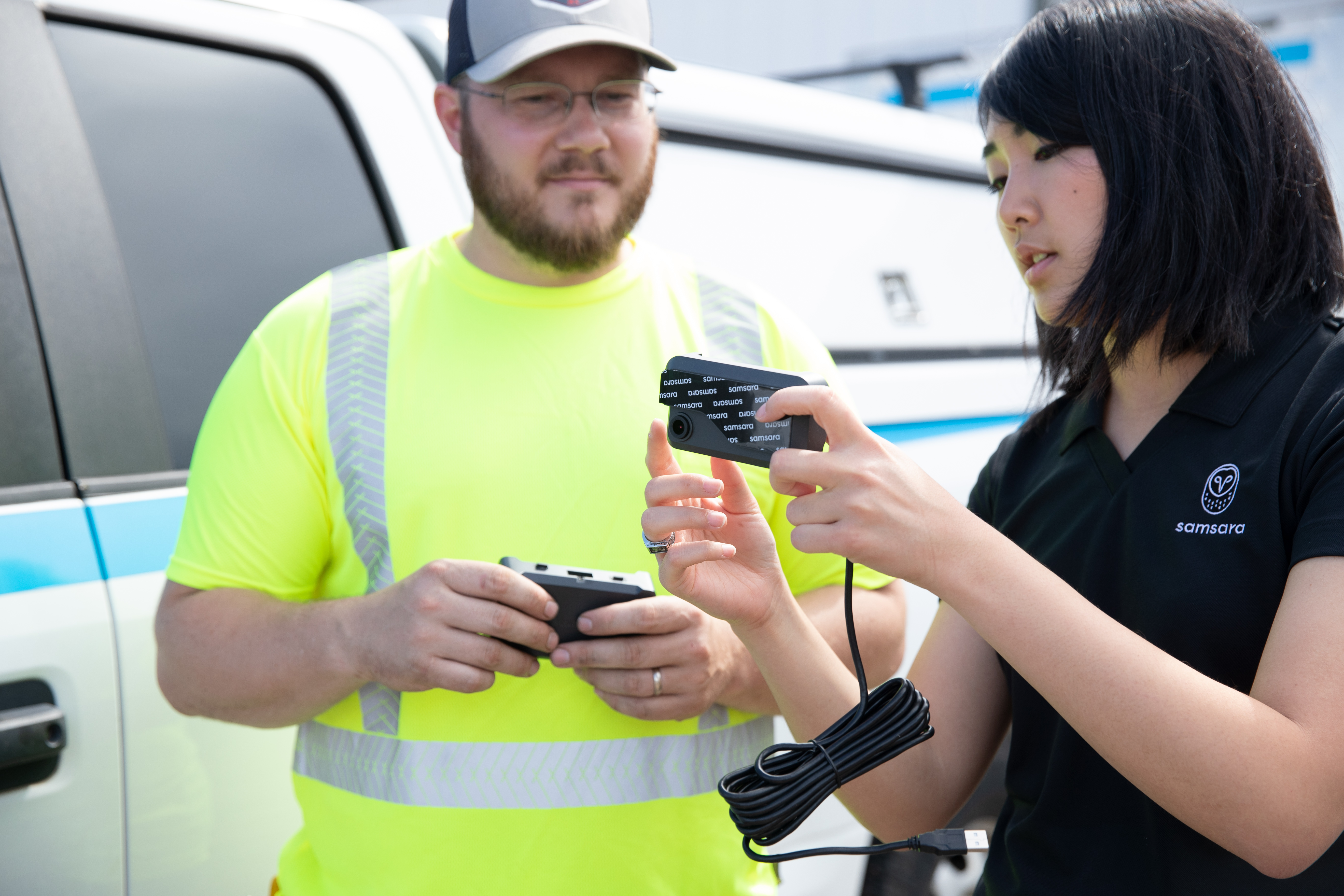 A Samsara rep in black shows a dashcam to a worker in a yellow safety vest near a white fleet vehicle.