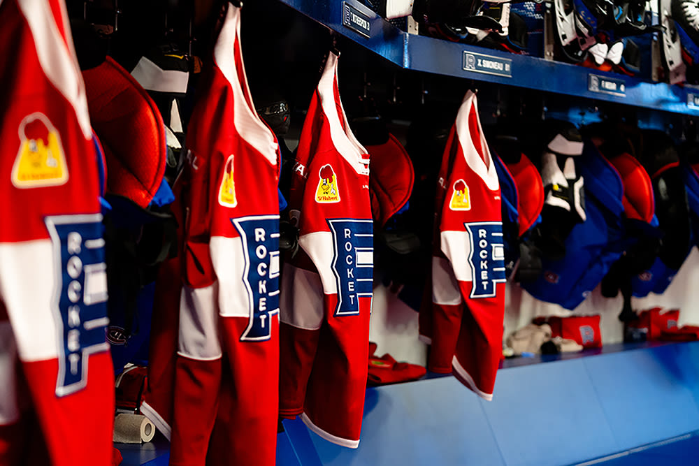 Red, white, and blue Laval Rocket hockey jerseys hanging in the locker room with yellow logo.