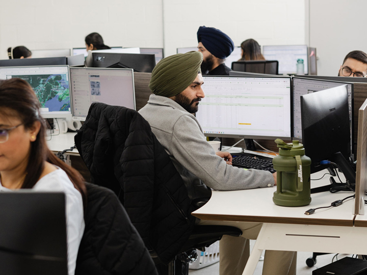 Dispatcher wearing a green turban and grey sweater looks at computer screens in office with colleagues.