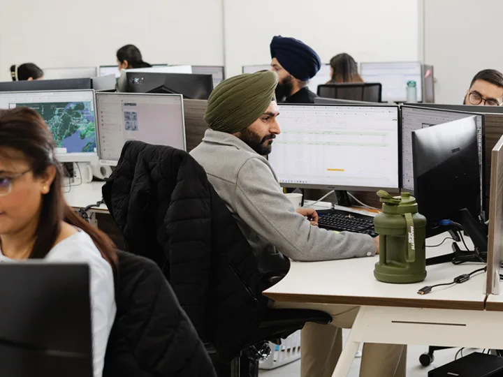 Dispatcher wearing a green turban and grey sweater looks at computer screens in office with colleagues.