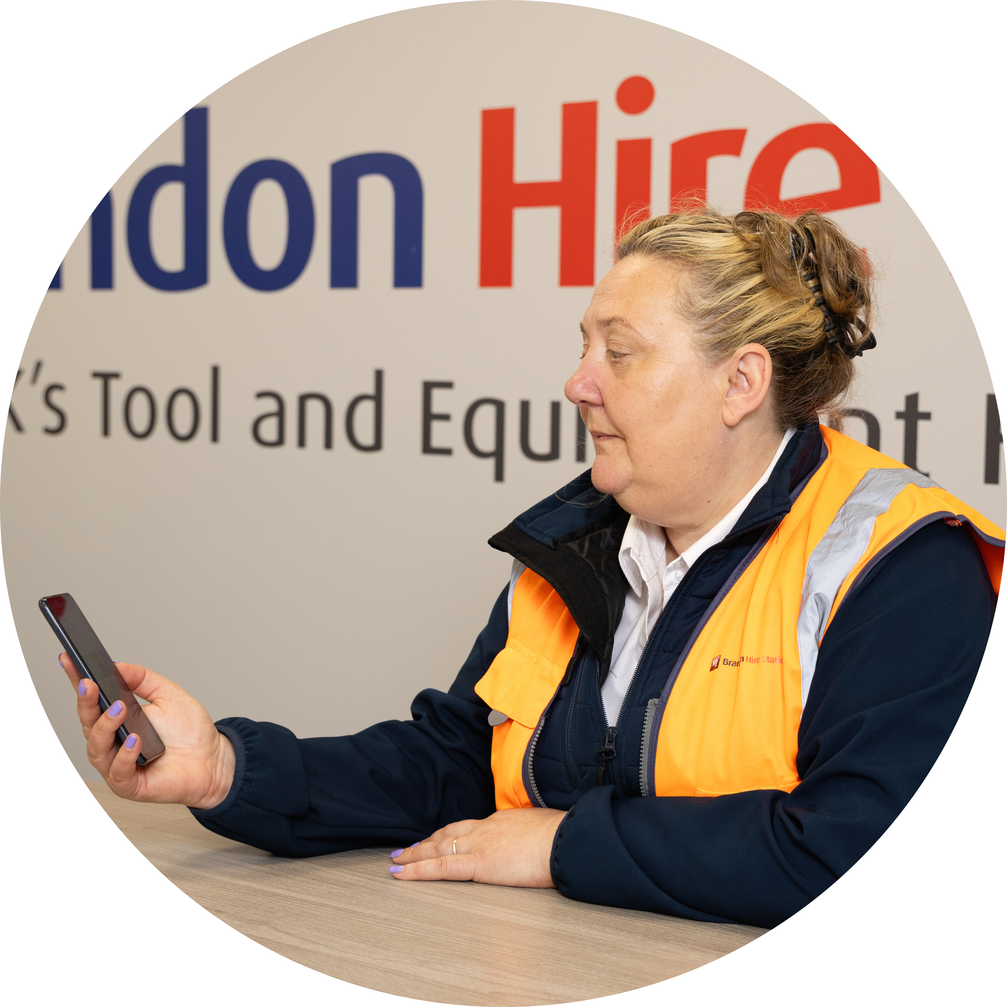 Person in orange safety vest checking phone at Brandon Hire counter, with company logo visible in background.