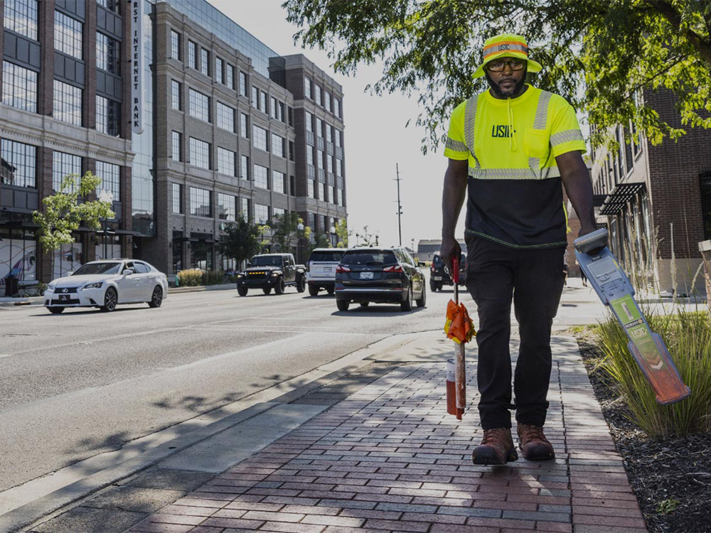 A worker in a yellow safety vest walks on a city sidewalk carrying equipment, with traffic and buildings in the background.