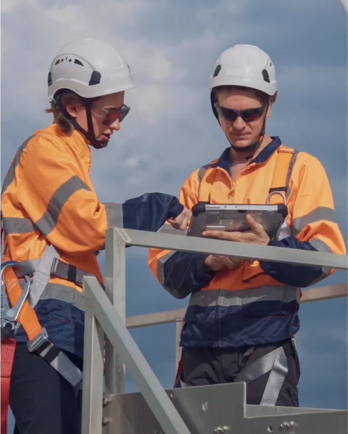 Two fleet managers in high-visibility safety gear and hard hats review tablet data at industrial site