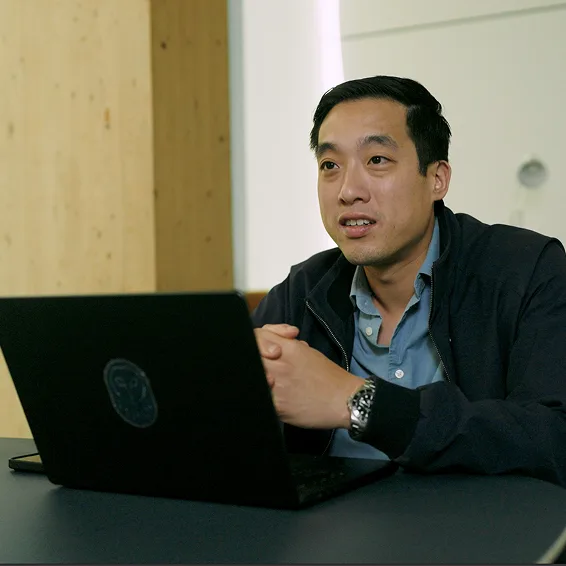 Person in dark jacket speaking while sitting at table with black laptop in modern wood-panelled room.