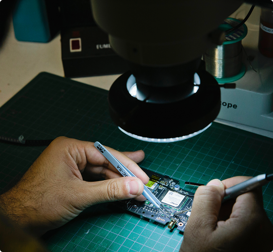 Hands repairing a circuit board under a microscope on a green cutting mat, using precision tools.