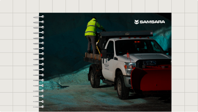 Worker in yellow vest stands on white truck with red snow plow at night, Samsara logo visible.