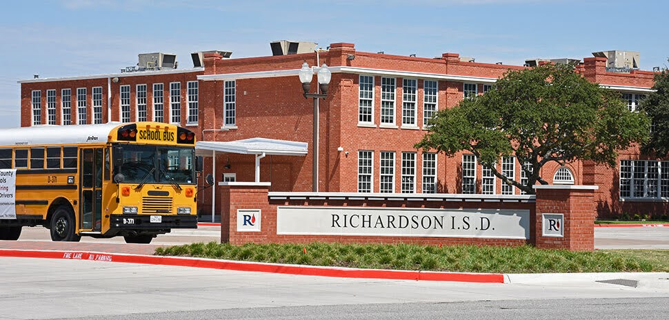 Yellow school bus parked outside a red brick Richardson ISD building with a district sign in the foreground.