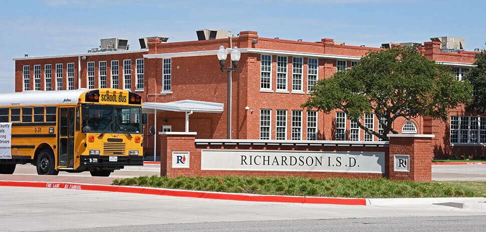 Yellow school bus parked outside a red brick Richardson ISD building with a district sign in the foreground.