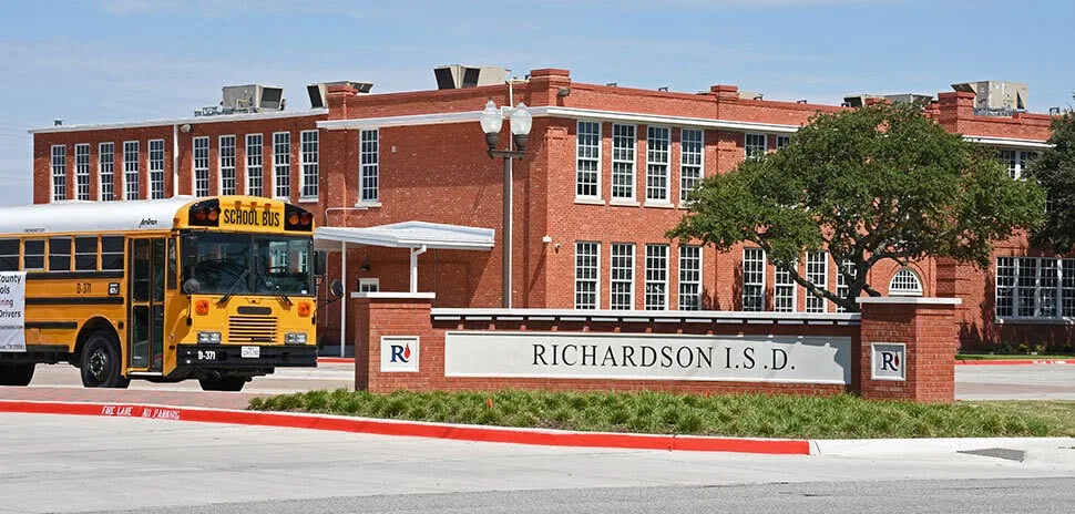 Yellow school bus parked outside a red brick Richardson ISD building with a district sign in the foreground.