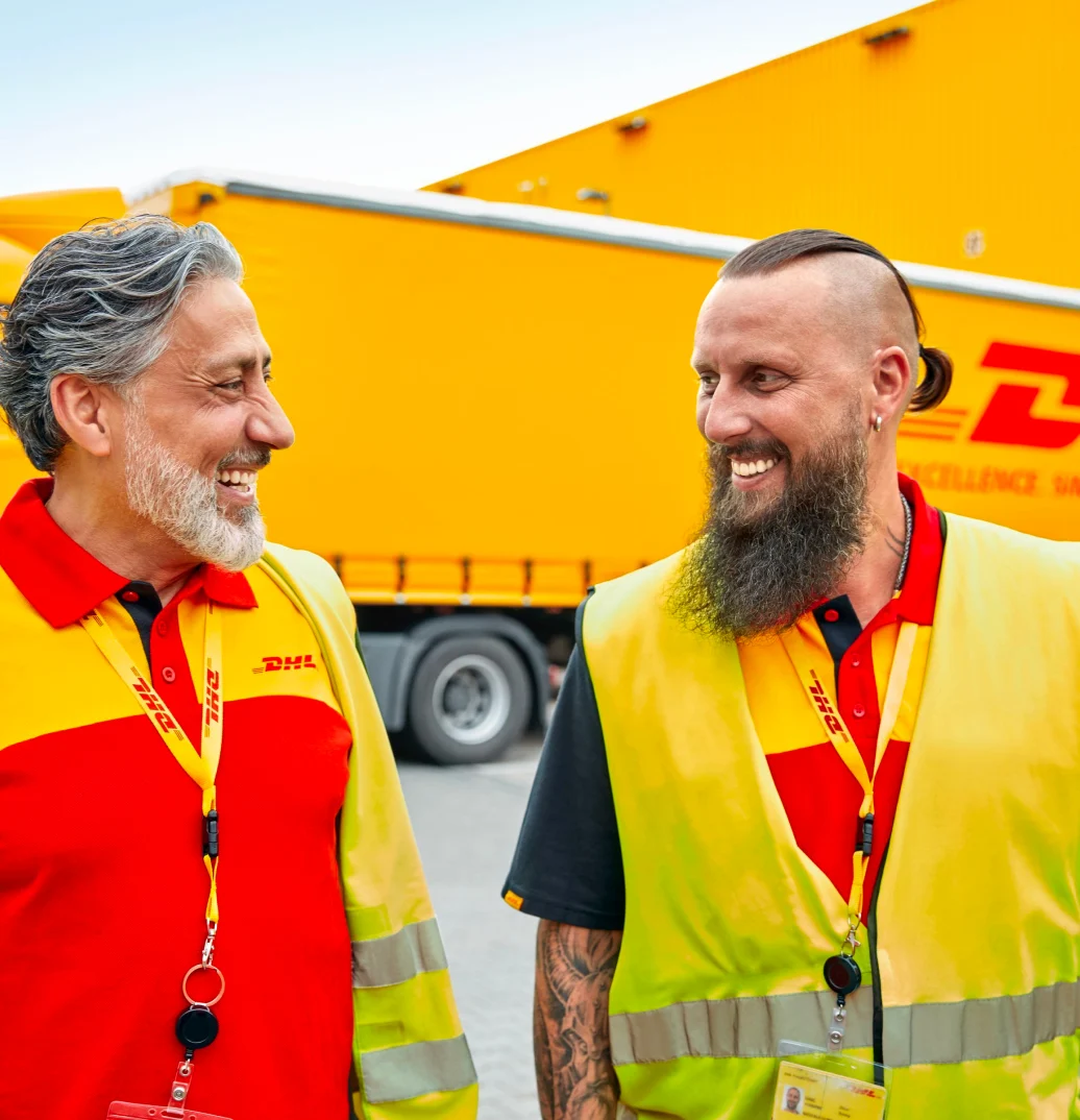 Two DHL employees looking at each other and smiling while standing in front of a DHL truck outside of a warehouse.