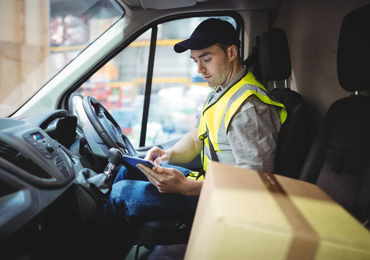 A delivery driver in a yellow hi-vis vest sits in a van, using a tablet, with a cardboard parcel on the seat beside him.