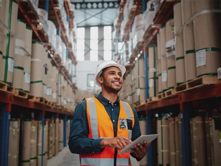Worker in safety vest and hard hat using tablet in large warehouse with tall shelving stacked with boxed inventory.