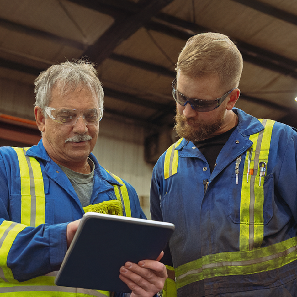 Two workers in blue safety uniforms with reflective stripes examining information on a tablet in an industrial setting.