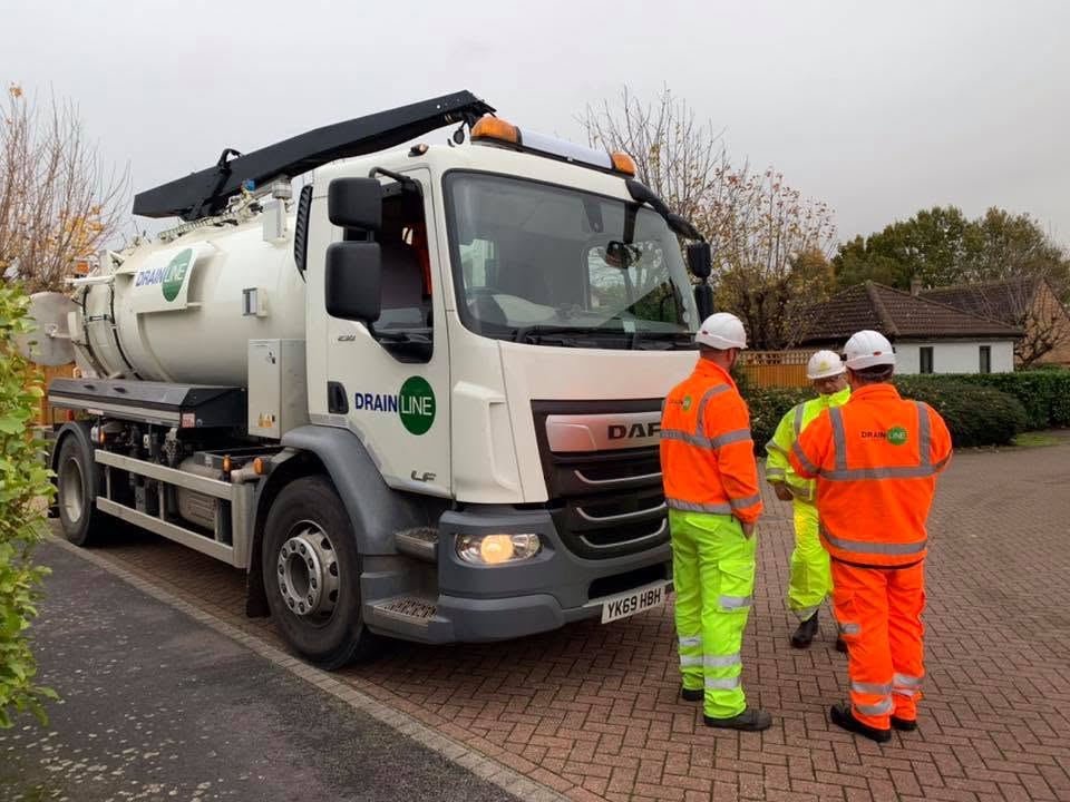 Three Drainline workers in hi-vis gear and hard hats stand beside a white Drainline DAF tanker truck on a residential street.