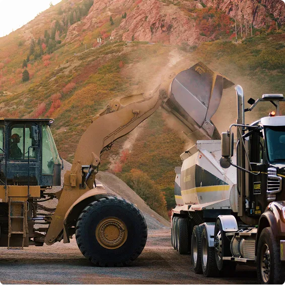 Front loader dumping material into a dump truck at a mountainside construction site with autumn foliage in background.