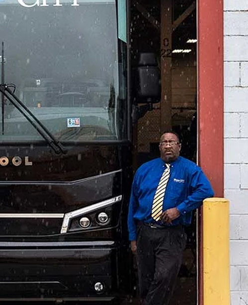 Bus driver in blue shirt with yellow tie standing in doorway of black coach bus next to white building.