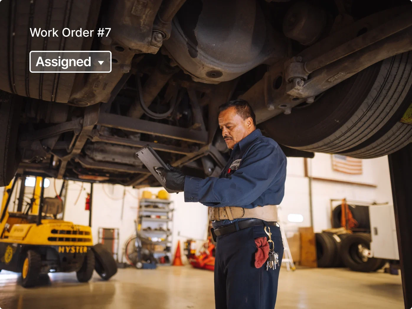 Mechanic in blue uniform checking vehicle undercarriage with tablet device, "Work Order #7" and "Assigned" status visible.