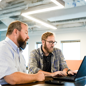 Two men collaborating at a desk, looking at a laptop in a bright office space with industrial ceiling.